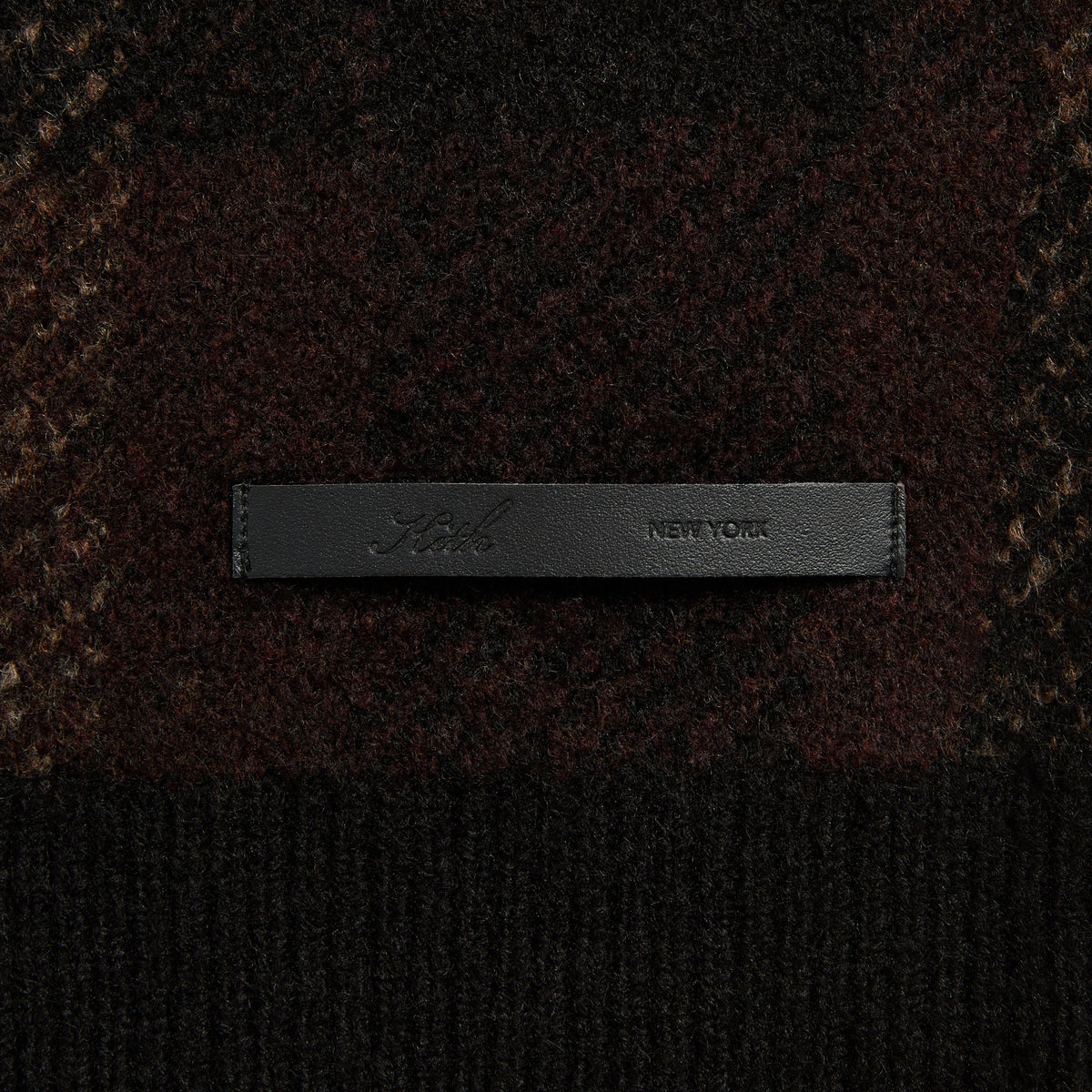 Close-up of the black leather 'Kith New York' label on the Lewis check sweater